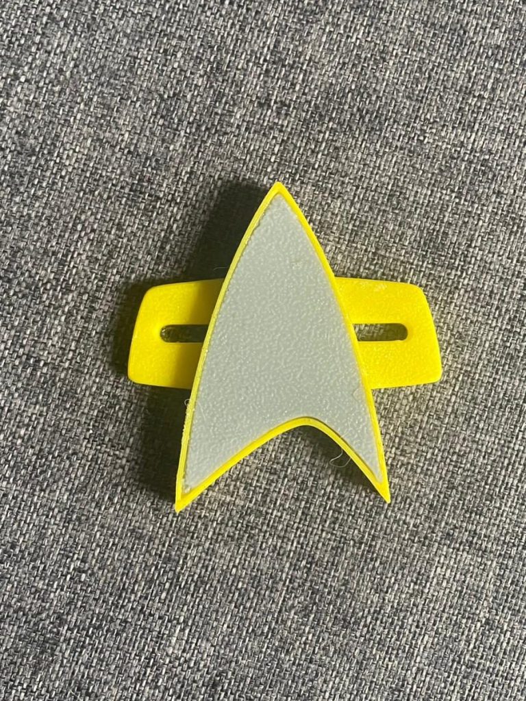 Gelbes Abzeichen mit einem stilisierten, grauen Sternenflottensymbol. 
